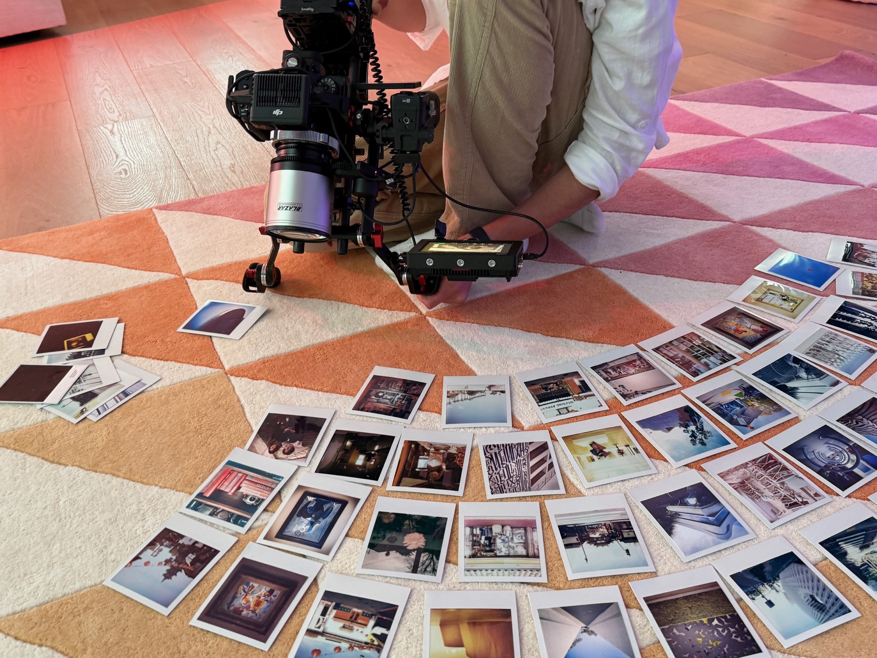 A camera on a low rig photographs dozens of Polaroid prints laid out across a patterned rug